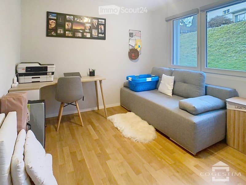 Bright living room with a grey sofa, white throw pillows, a small wooden table with a printer, a grey chair, a blue basket on the sofa, and a white shag rug on the wooden floor. Large windows offer a view of greenery outside.