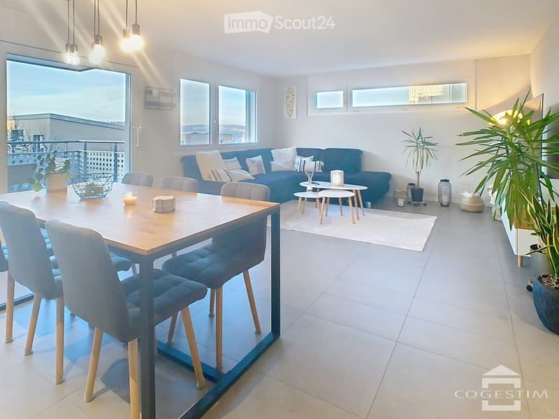 Bright, modern living and dining area with large windows offering a city view. Features a blue sofa, wooden dining table with chairs, small coffee tables, and decorative plants.