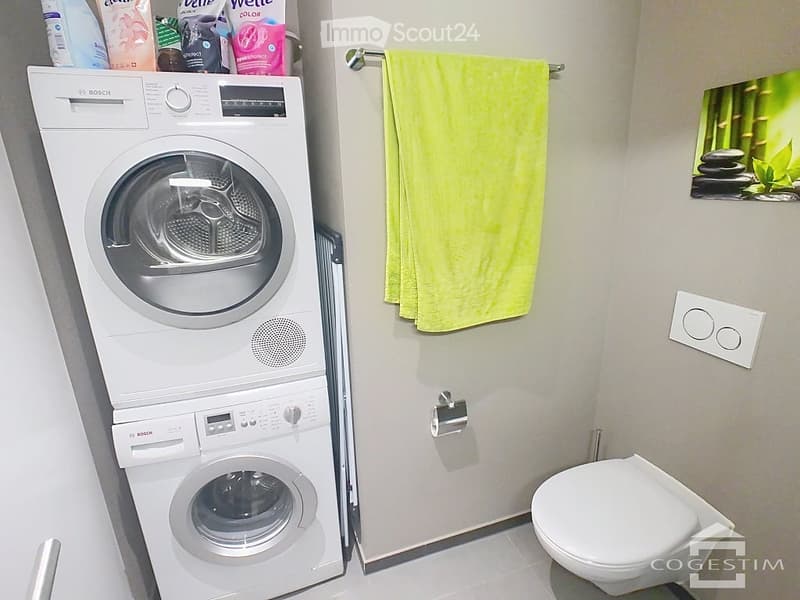 Modern bathroom with a stacked Bosch washing machine and dryer, a toilet, a yellow towel hanging on a rack, and a decorative picture on the wall.