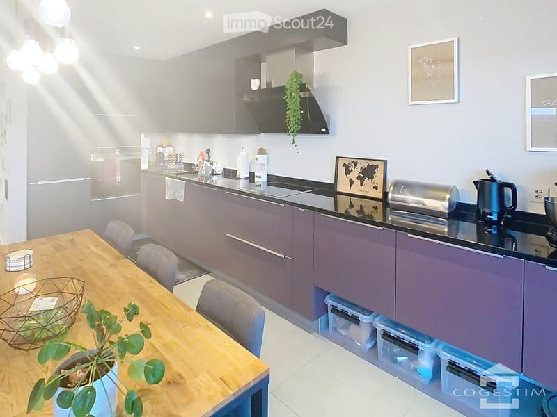 Modern kitchen with grey cabinets, black countertops, and stainless steel appliances. Features a wooden dining table with chairs, a potted plant, and decorative items on the counter.