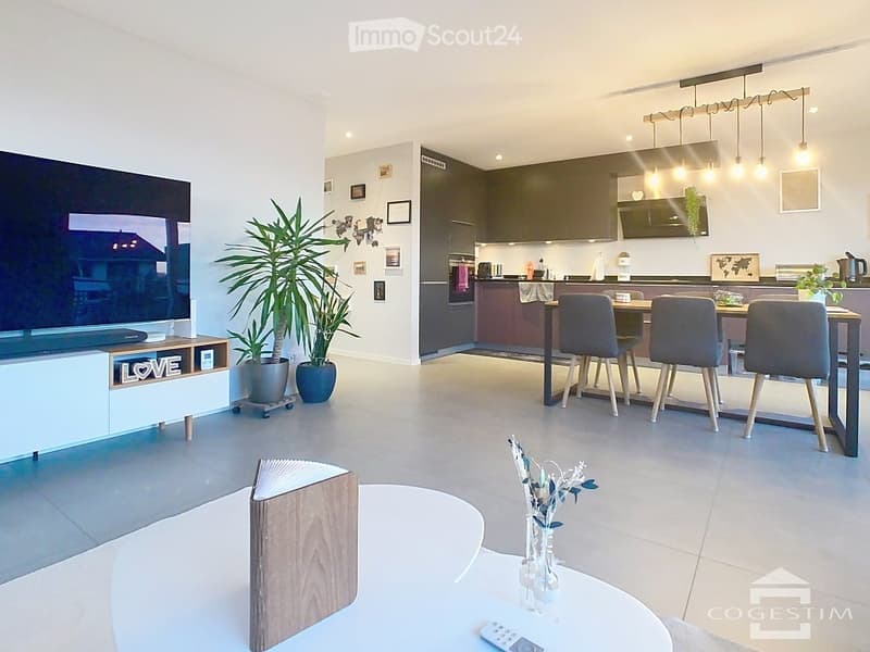 Open-plan living and dining area with modern furniture, including a sofa, coffee table, TV stand, dining table with chairs, and kitchen cabinets. Potted plants and decorative items are present.