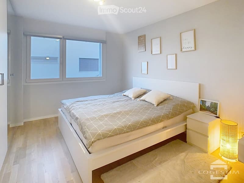 Modern bedroom with a white bed, grey bedding, white nightstand, and decorative picture frames on the wall. The room features large windows and light wooden flooring.