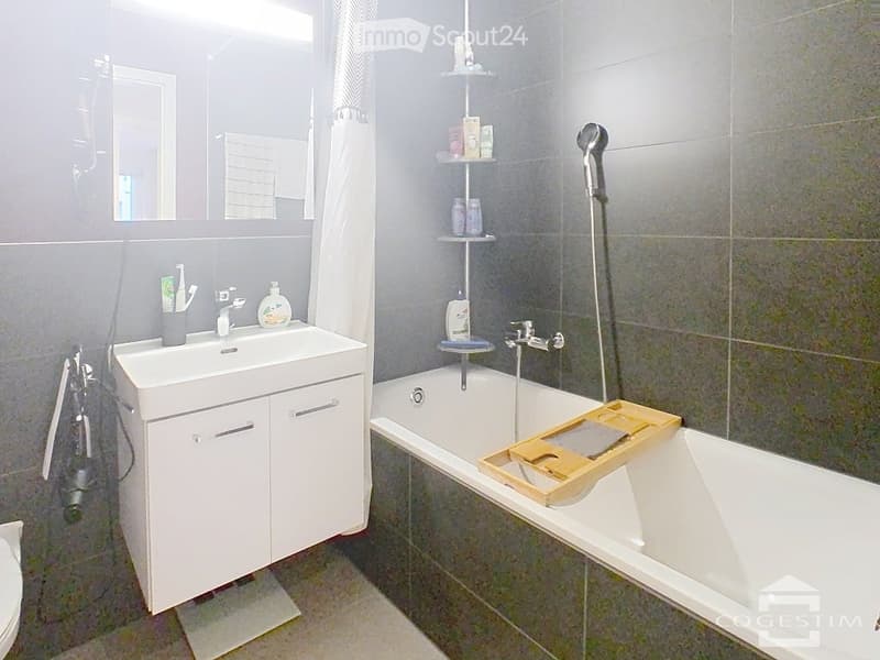 Modern bathroom with a white sink, bathtub, and mirrored wall. Includes a wooden tray on the edge of the tub and a small shelf for toiletries.