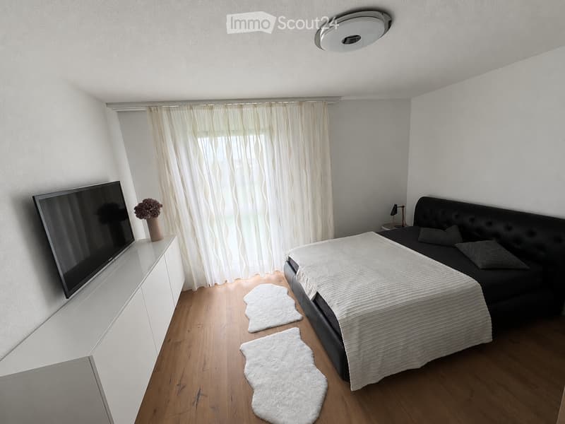Modern living room with a black leather sofa, a white throw blanket, a flat-screen TV on a white stand, a small table with a vase of flowers, and two white rugs on the wooden floor.