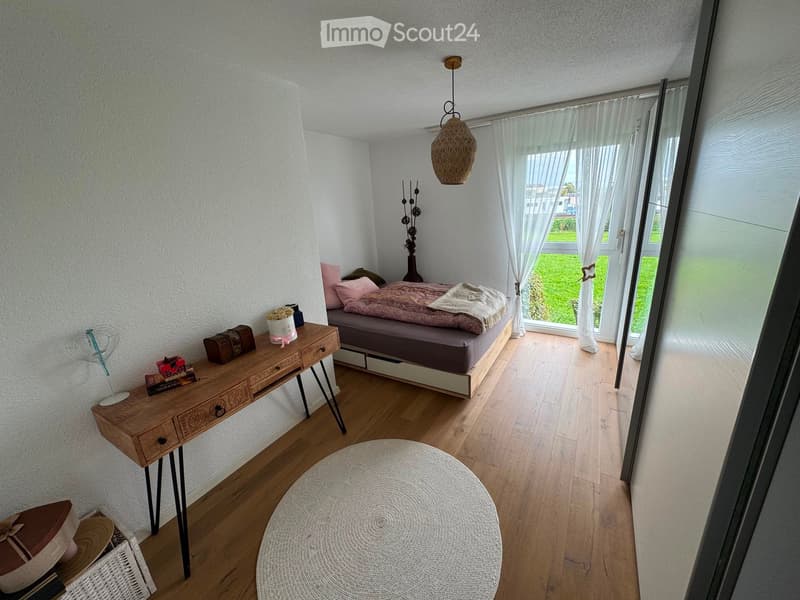 Cozy bedroom with wooden flooring, a bed with pink bedding, a nightstand with a vase, a small table with books and a fan, and a round white rug.