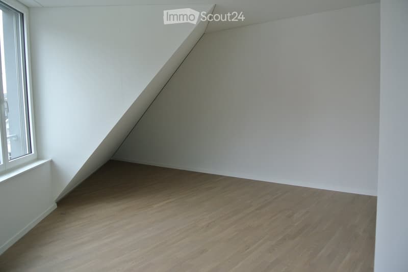 Empty room with slanted ceiling, wooden floor, and white walls. There is a large window on the left side of the room.