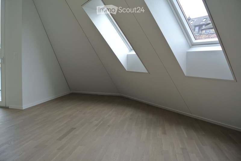 An empty room with wooden flooring, white walls, and two triangular windows. The ceiling is slanted, providing a view outside.