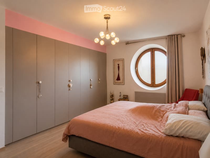 Modern bedroom with a large bed, wardrobe, round window, chandelier, and a small table with a picture frame.