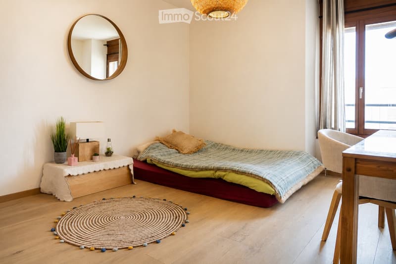 Minimalist bedroom with a low platform bed, a round mirror, a small table with a lamp and plants, and a woven circular rug on the floor.