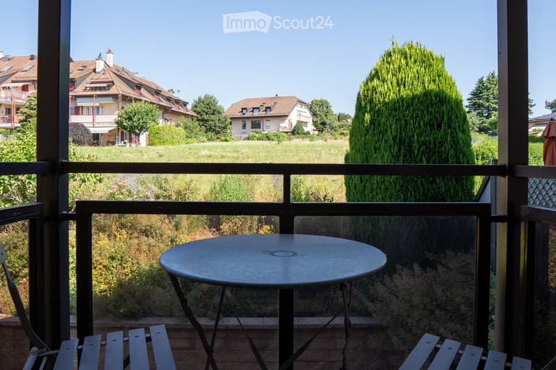 Balcony with a round metal table and chairs, overlooking a grassy area with trees and houses in the background.
