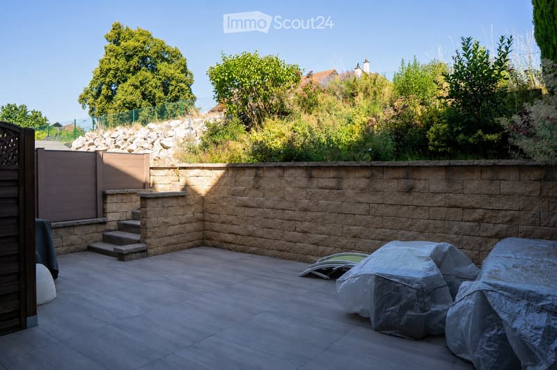 A well-maintained garden area with a stone wall, steps leading up, covered furniture, and lush greenery.