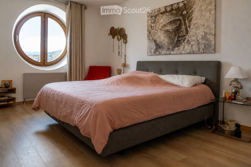 Modern bedroom with a large bed, nightstand, red armchair, round window, and artwork on the wall.