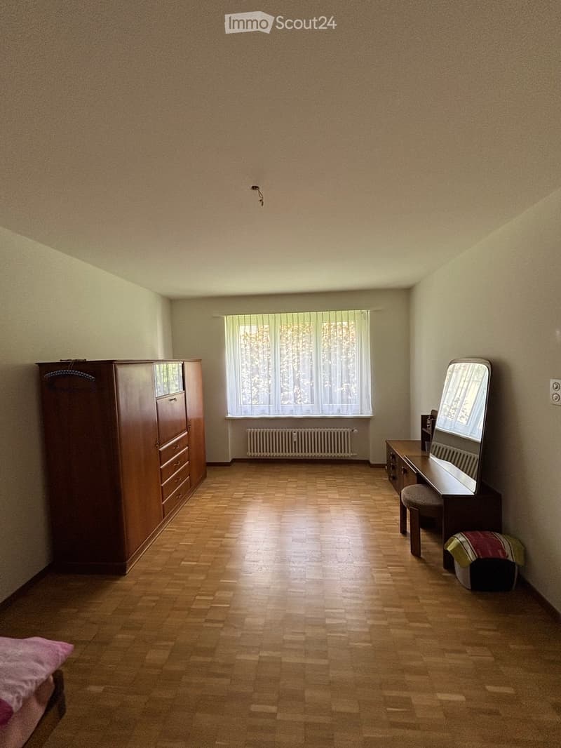 A furnished room with wooden flooring, a large wardrobe, a mirror, a small table, and a radiator under the window.