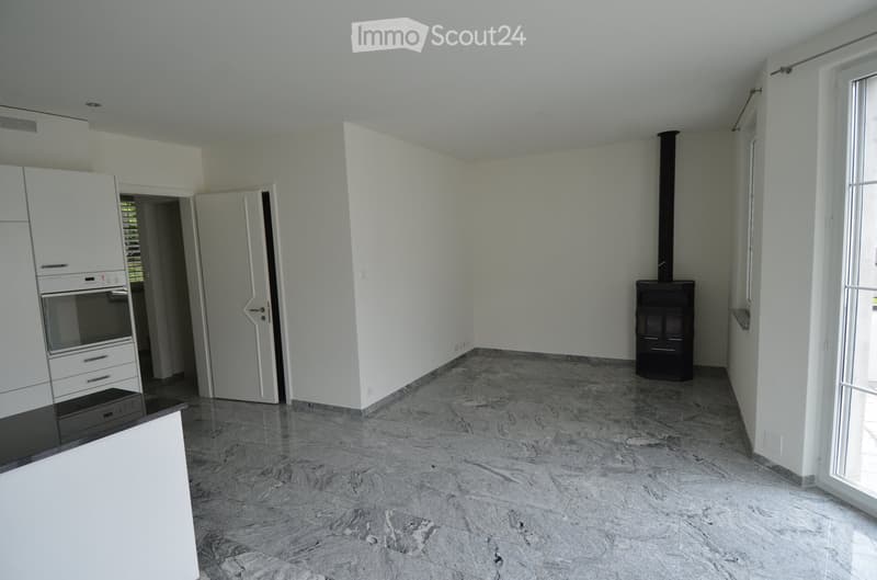 Empty room with white walls, marble flooring, and a fireplace in the corner. There is a kitchen area with cabinets and an oven visible through an open doorway.