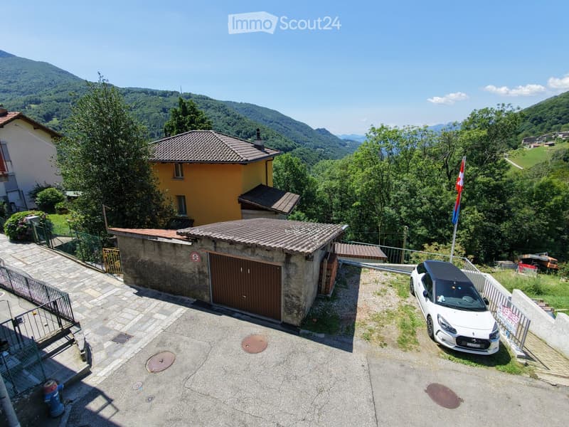 Aerial view of a residential area with a stone garage, a yellow house, and a white car parked on a paved driveway, surrounded by greenery and mountains.