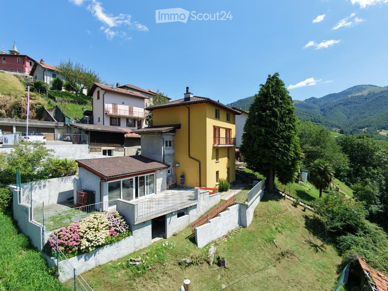 A multi-story residential house with a yellow facade, red shutters, and a tiled roof, situated on a hillside with a well-maintained garden and surrounding greenery.