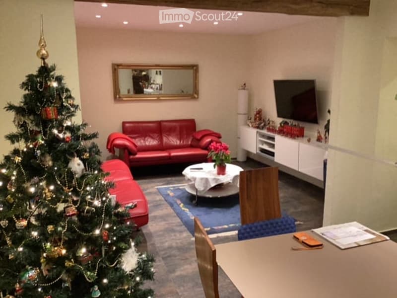 A cozy living room with a red leather sofa, a coffee table, a mirror above it, a television on a white cabinet, and a Christmas tree on the left side.