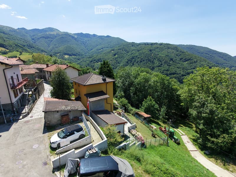 Aerial view of a rural village with houses, a parked car, and surrounding greenery and mountains.