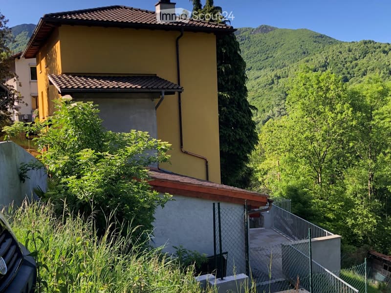 Two-story yellow house with a tiled roof, surrounded by greenery and a mountainous backdrop. A staircase leads up to the entrance.