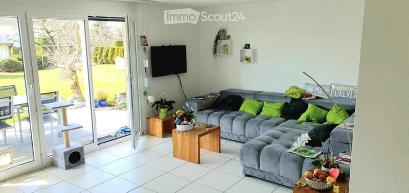 Bright living room with large sliding glass doors opening to a garden, grey sectional sofa with green cushions, wooden coffee table, cat tree, cat house, small table with flower pot, and a television mounted on the wall.