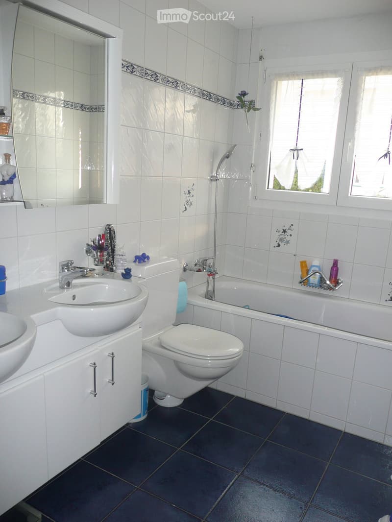 Modern bathroom with white tiles, dark blue floor tiles, a sink, toilet, bathtub, and a mirror. The room features a window with a white curtain and decorative tiles on the wall.