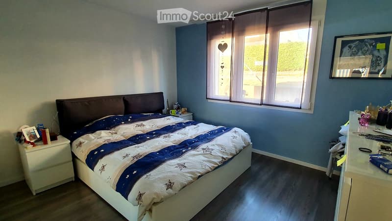 Cozy bedroom with a large bed, nightstand, desk, and a large window with blinds. The room features a blue and white star-patterned duvet and a wooden floor.