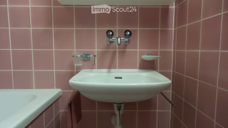 A small bathroom with pink tiled walls and a white sink, faucet, toothbrush holder, toothpaste holder, and bathtub.