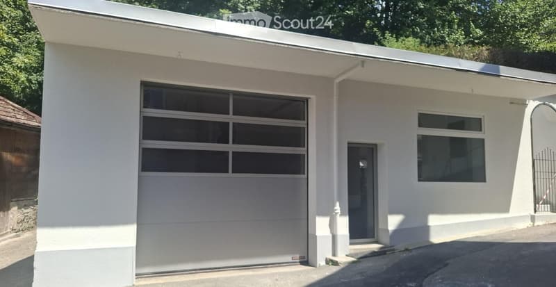 Modern single-story building with a flat roof, large garage door, and small entrance door. The exterior is painted white with clean lines and large windows.