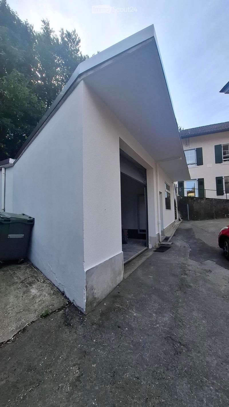 A modern, white building with a flat roof and a covered entrance area. The entrance leads to a small porch with a concrete floor. A red car is partially visible on the right side, and a green trash bin is on the left.