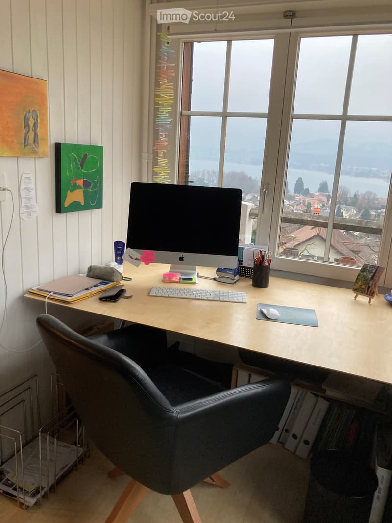 A well-organized home office with a large wooden desk, a black leather chair, a computer setup, and various office supplies. The room features large windows with scenic views and is decorated with colorful artworks.