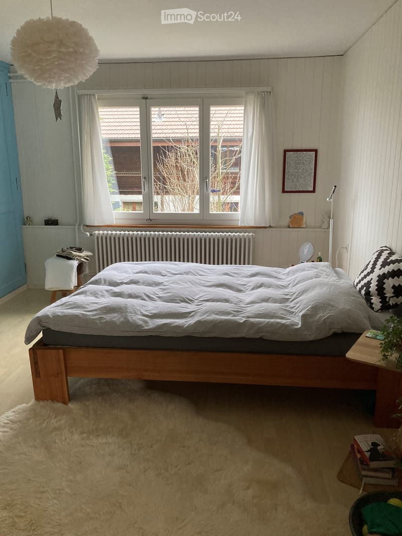 Cozy bedroom with a wooden bed, nightstand, mirror, radiator, stool, picture frame, books, plant, and a decorative ball lamp.