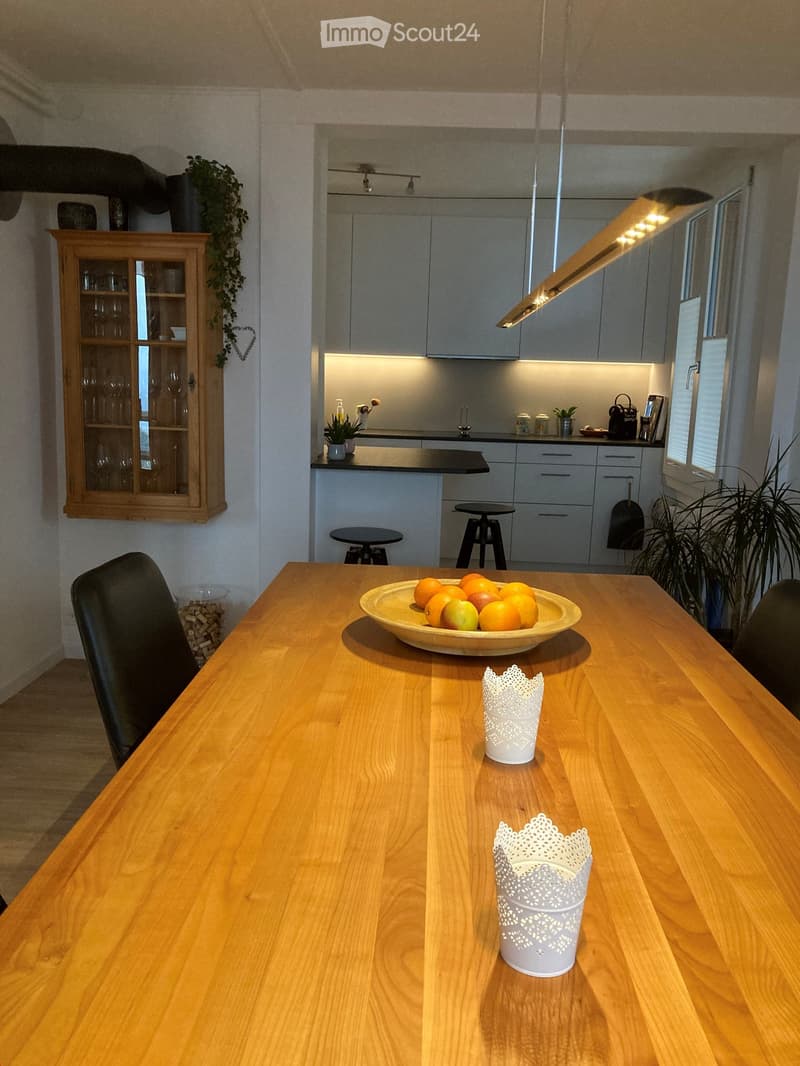 Modern kitchen with grey cabinets, wooden dining table, and various decorative items including a bowl of fruit and two white decorative cups.