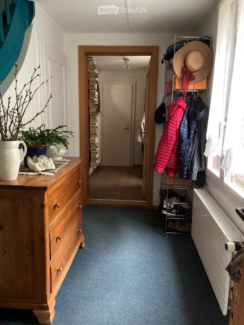 Hallway with blue carpet, wooden dresser, coat rack, and mirror leading to another room.