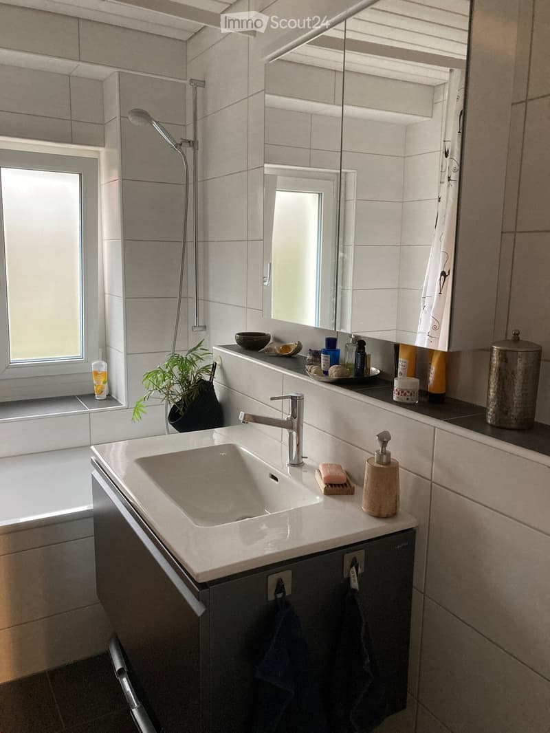 Modern bathroom with a white sink, chrome faucet, large mirror, shower area, and various toiletries on the counter.