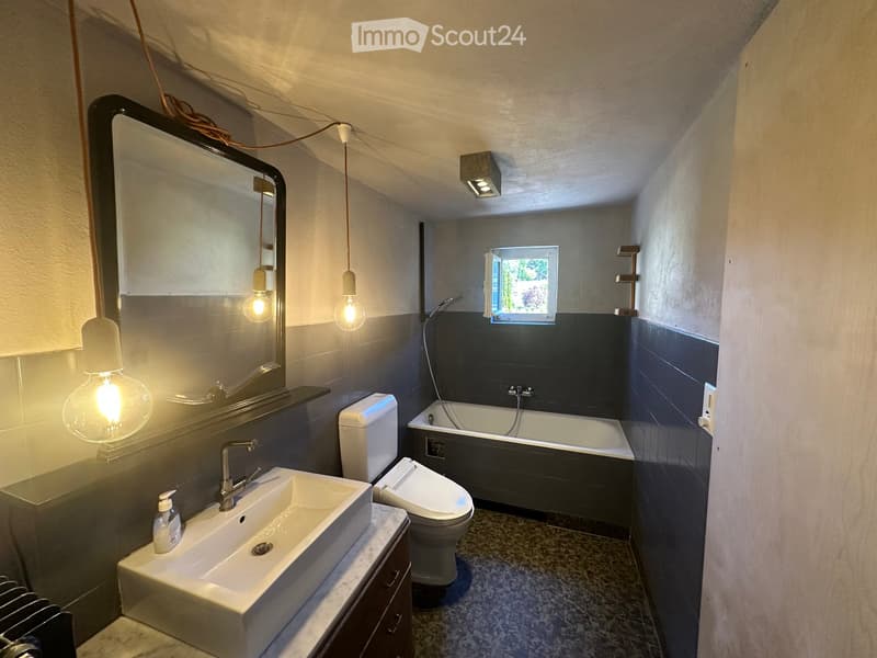 Modern bathroom with a white sink, toilet, bathtub, and large mirror. Features pendant lights and a window above the bathtub.