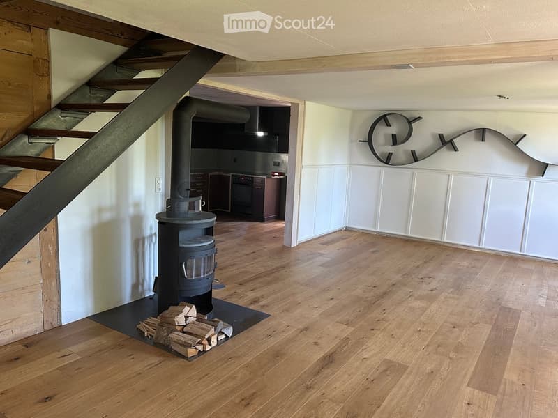 Spacious room with wooden flooring, a central fireplace with stacked firewood, and a modern wall-mounted clock. A staircase leads upwards, and a kitchen area is visible in the background.