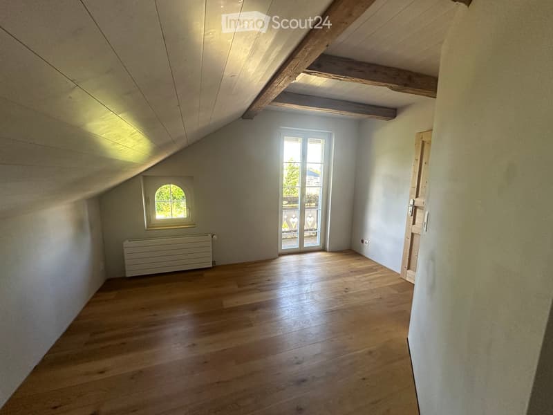 Empty attic room with wooden flooring, slanted wooden ceiling beams, white walls, and two windows, one of which opens to a balcony.