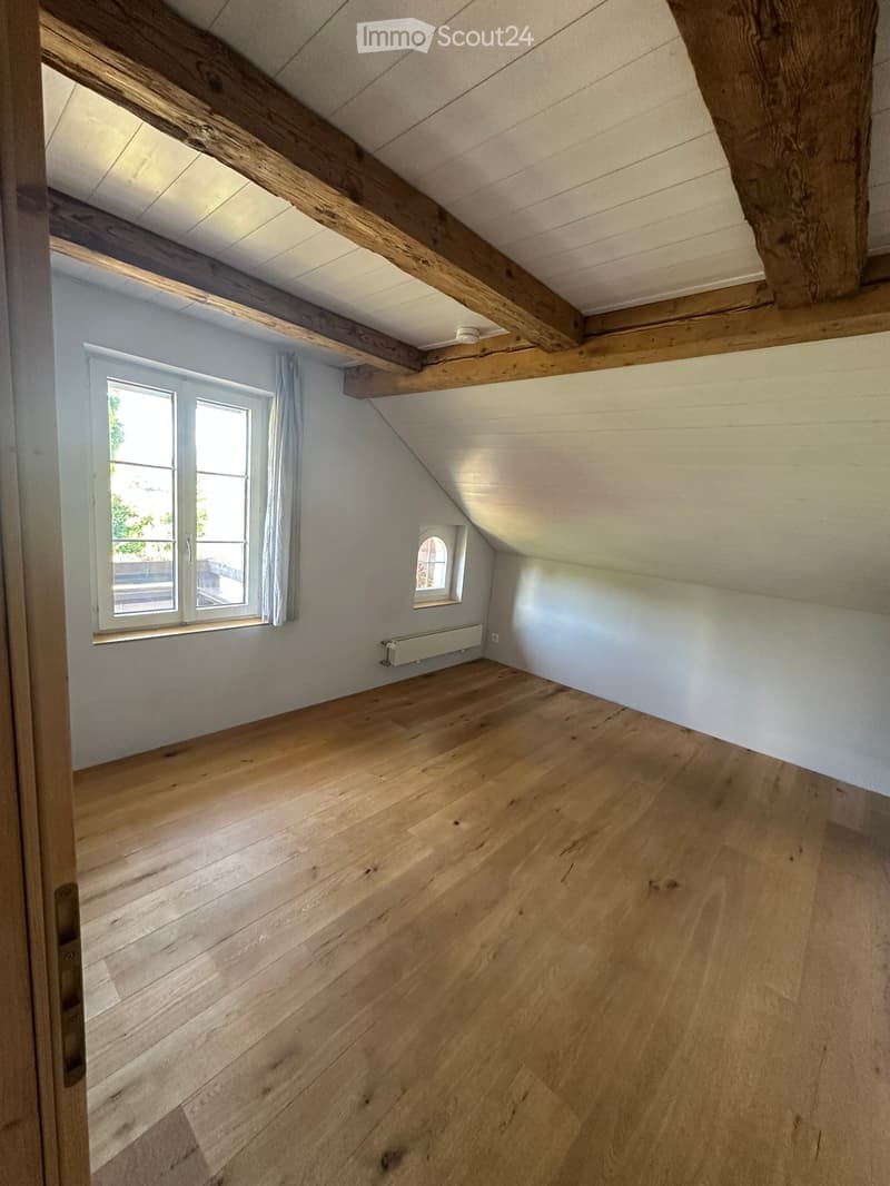 Empty attic room with wooden floors, exposed wooden beams, white walls, and two windows, one with a curtain.