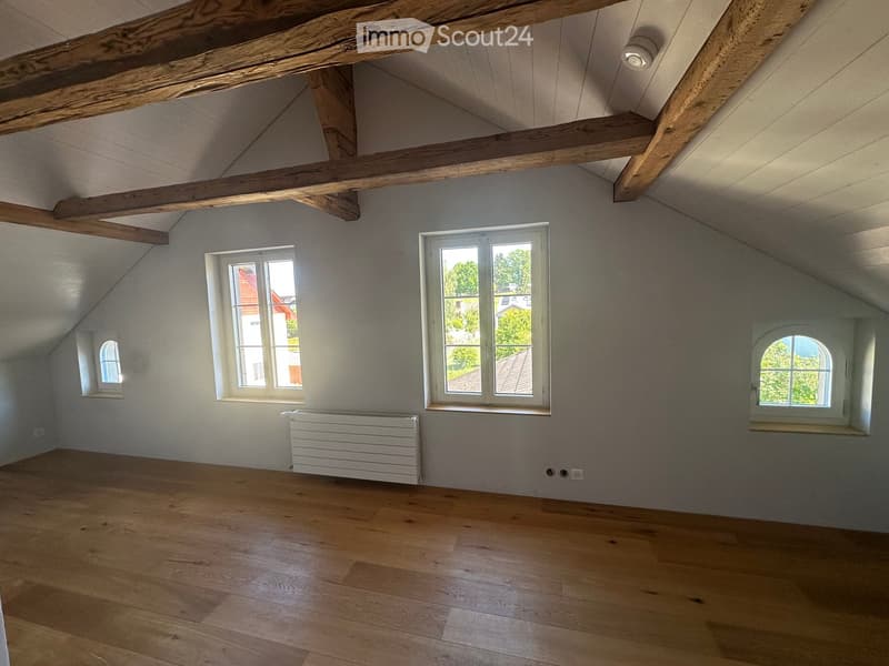 Empty attic room with wooden beams, wooden flooring, and multiple windows offering natural light.