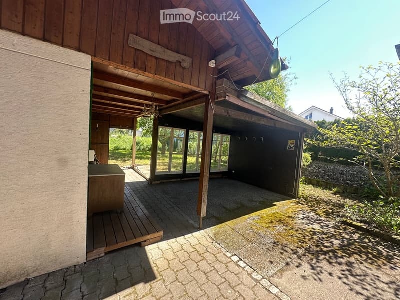 Wooden covered outdoor area with a garage, wooden bench, and surrounding greenery.