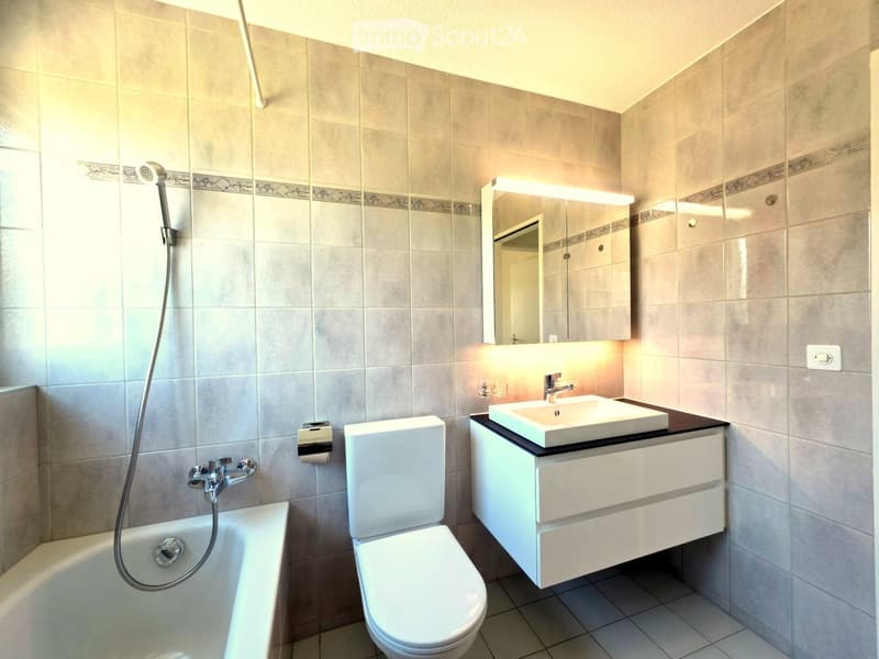 Modern bathroom with beige tiles, a bathtub, toilet, sink with a mirrored cabinet, and a showerhead.