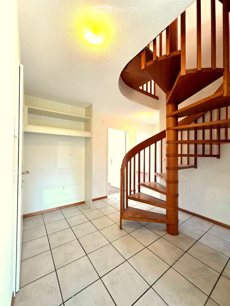 Spacious hallway with a wooden spiral staircase, tiled floor, and white walls. Features a built-in shelf unit and a door leading to another room.
