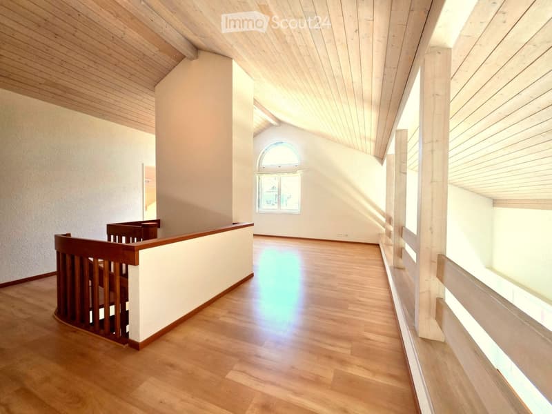 Bright, open hallway with wooden flooring and white walls, featuring a wooden railing and large windows allowing natural light to flood the space.
