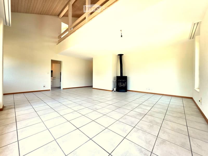 Empty room with white walls, tiled floor, and a black wood-burning stove with a chimney. The room has wooden trim and a slanted wooden ceiling with exposed beams.