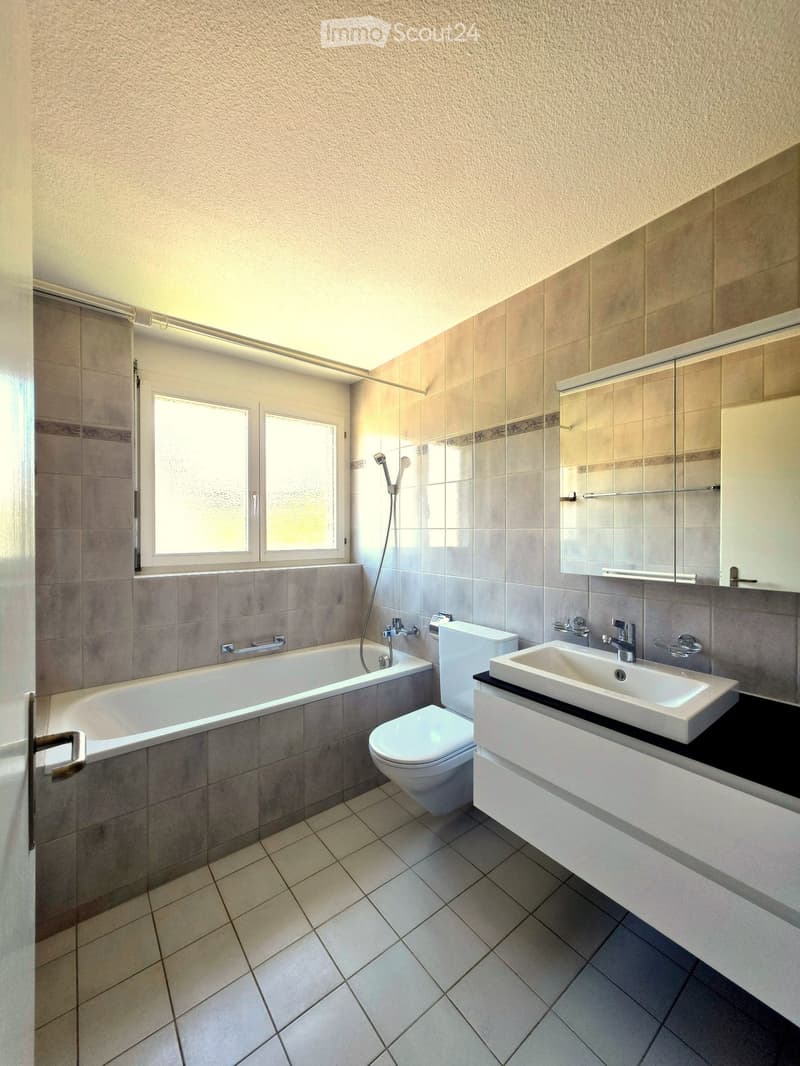 Modern bathroom with a bathtub, toilet, sink, mirror, and showerhead. Features tiled walls and floor, a window, and a built-in vanity with a sink.