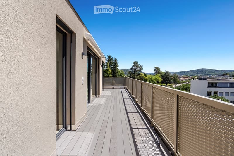 Balcony with wooden decking, metal railing, and glass doors leading to the interior. The view includes trees and distant hills.