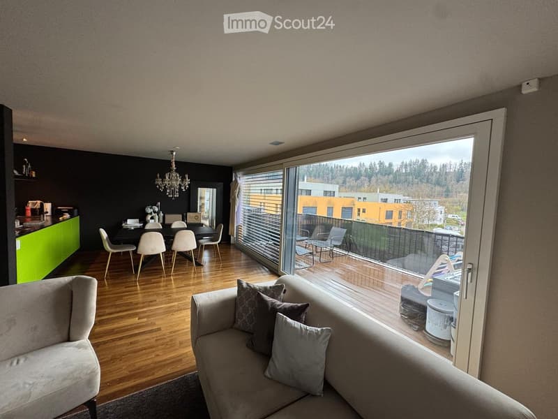 Modern living and dining area with a grey sofa, coffee table, dining table with chairs, and a kitchen counter. Large windows open to a balcony with outdoor seating.