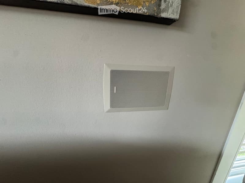 An unfurnished room with a white wall-mounted speaker and a painting on the wall above it.