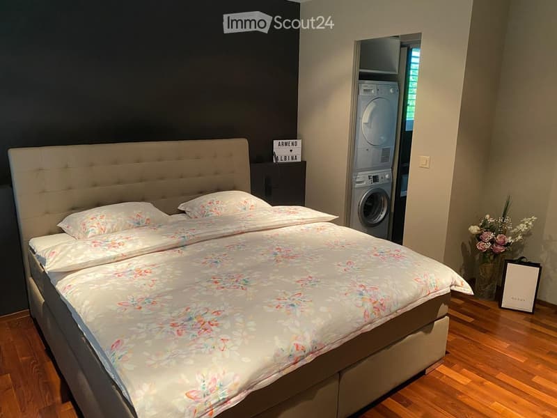 Bedroom with a large double bed, floral bedding, a private washing machine, and a vase with flowers on the floor.