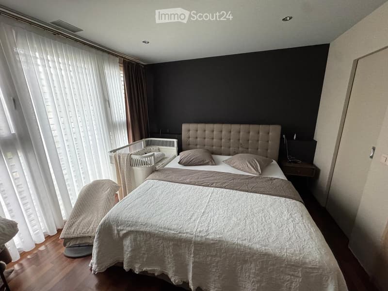 A well-lit bedroom with a large bed, a crib, and a nightstand. The room features wooden flooring and large windows with white curtains.